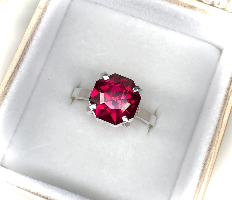 May include: A silver ring with a large, faceted, ruby-red gemstone. The ring is set in a simple, modern style, and is presented in a white jewelry box with a soft, velvety interior.