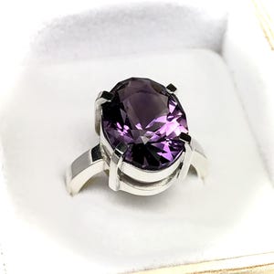 May include: A silver ring featuring a large, oval-cut, deep purple gemstone. The gemstone is set in a four-prong setting, and the band is a simple, polished silver. The ring is displayed in a white jewelry box.
