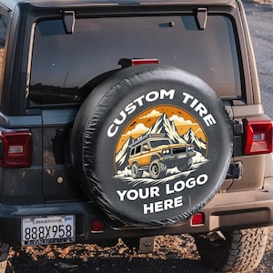 Può includere: Un SUV Jeep nero con una copertura per pneumatici di scorta con una grafica di montagna e veicolo. La copertura recita "CUSTOM TIRE" e "YOUR LOGO HERE". Una persona è seduta sul tetto del veicolo, affacciata su un paesaggio montano.