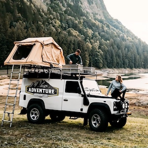 Puede incluir: Un veh&iacute;culo todoterreno blanco con una tienda de campa&ntilde;a en el techo, una escalera y las palabras "Adventure Outdoor Car" en el lateral. Dos personas est&aacute;n de pie cerca del veh&iacute;culo.