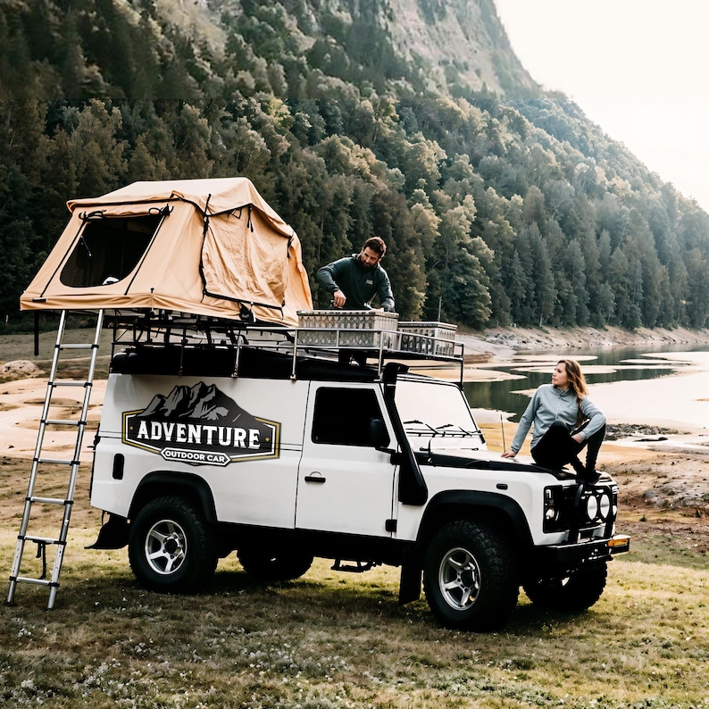 Puede incluir: Un veh&iacute;culo todoterreno blanco con una tienda de campa&ntilde;a en el techo, una escalera y las palabras "Adventure Outdoor Car" en el lateral. Dos personas est&aacute;n de pie cerca del veh&iacute;culo.