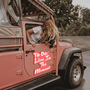 Puede incluir: Un Jeep rosa con un letrero rosa y blanco que dice "To Do: Live In The Moment". Una mujer con el pelo largo y rubio se asoma por la ventana del lado del conductor.
