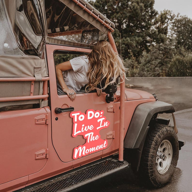 Puede incluir: Un Jeep rosa con un letrero rosa y blanco que dice "To Do: Live In The Moment". Una mujer con el pelo largo y rubio se asoma por la ventana del lado del conductor.
