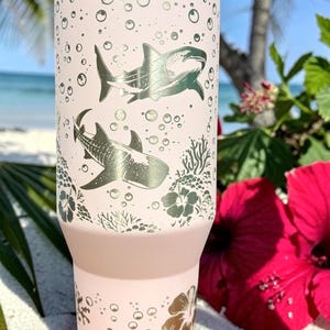 May include: A light pink insulated tumbler featuring silver shark and bubble designs. The tumbler has a tapered base and a wide mouth. Floral and coral designs are at the bottom. The background includes a beach scene with red flowers.