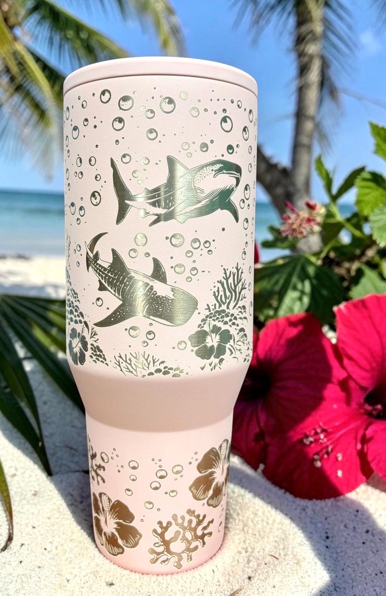 May include: A light pink insulated tumbler featuring silver shark and bubble designs. The tumbler has a tapered base and a wide mouth. Floral and coral designs are at the bottom. The background includes a beach scene with red flowers.