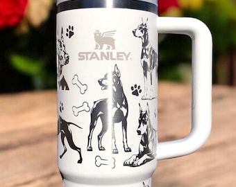 Può includere: Un bicchiere Stanley bianco con manico e cannuccia, caratterizzato da un motivo grafico nero con cani Dobermann, impronte di zampe e ossa. Il marchio Stanley è stampato in nero sulla parte superiore del corpo cilindrico di colore bianco.