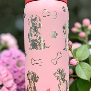 May include: Pink Owala water bottle with a red lid, featuring illustrations of dachshunds, paw prints, and dog bones. The bottle has a handle and a flip-top lid. The brand name "owala" is printed on the bottle.