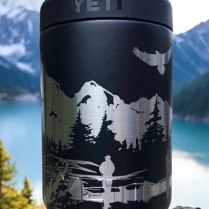 K&ouml;nnte beinhalten: Schwarzer YETI-Becher mit silbernem Berg- und Waldmotiv. Das Design zeigt einen fliegenden Adler und eine Person in der N&auml;he eines Sees. Der zylindrische Becher steht auf einem Felsen.