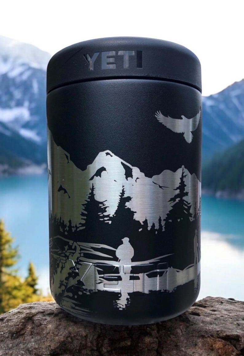 K&ouml;nnte beinhalten: Schwarzer YETI-Becher mit silbernem Berg- und Waldmotiv. Das Design zeigt einen fliegenden Adler und eine Person in der N&auml;he eines Sees. Der zylindrische Becher steht auf einem Felsen.