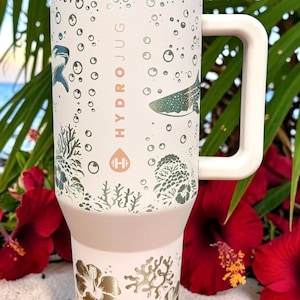 May include: A light pink HydroJug water bottle with a white handle and lid. The bottle features a design of marine life, including sharks and coral, with the word "HYDROJUG" printed vertically. The bottom of the bottle has a gold floral design.