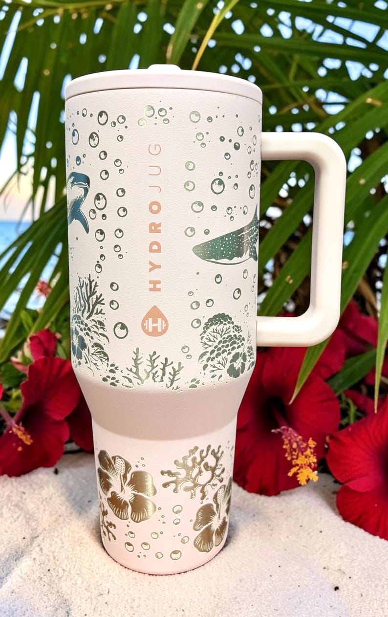 May include: A light pink HydroJug water bottle with a white handle and lid. The bottle features a design of marine life, including sharks and coral, with the word "HYDROJUG" printed vertically. The bottom of the bottle has a gold floral design.