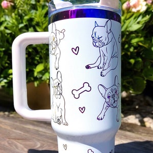 May include: White tumbler with a handle, featuring line drawings of French Bulldogs, hearts, and bones. The tumbler has a clear lid and a purple rim. The background shows flowers and sunlight.