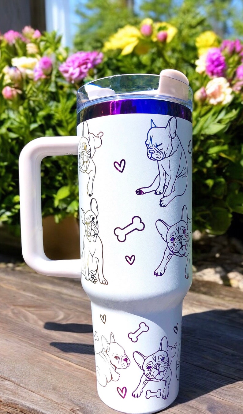 May include: White tumbler with a handle, featuring line drawings of French Bulldogs, hearts, and bones. The tumbler has a clear lid and a purple rim. The background shows flowers and sunlight.