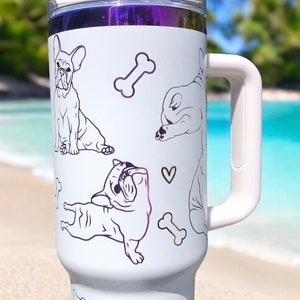 May include: White travel mug with a handle, featuring line art illustrations of French Bulldogs and dog bones. The mug has a purple metallic band at the top and a white handle. The background is a beach scene.