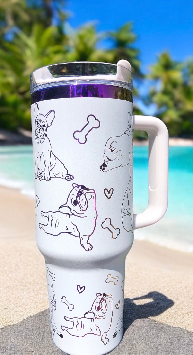 May include: White travel mug with a handle, featuring line art illustrations of French Bulldogs and dog bones. The mug has a purple metallic band at the top and a white handle. The background is a beach scene.