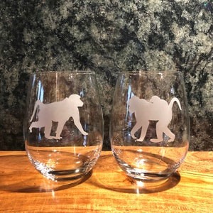 May include: Two stemless wine glasses with etched baboon silhouettes. The clear glasses sit on a wooden surface against a dark, textured background. The baboon design is a light, frosted color.