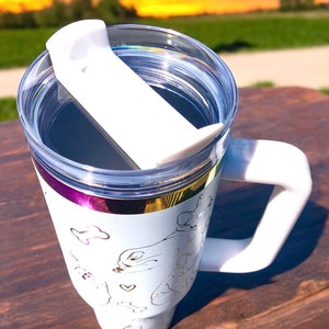 May include: White travel mug with a clear lid and a white handle. The mug features a gold and purple band around the top and a gold outline design of dogs and bones. The lid has a white slider to close the opening.