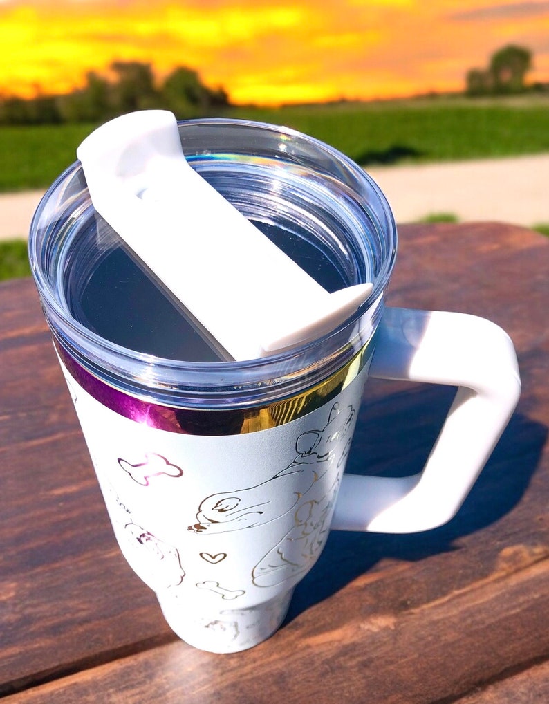 May include: White travel mug with a clear lid and a white handle. The mug features a gold and purple band around the top and a gold outline design of dogs and bones. The lid has a white slider to close the opening.