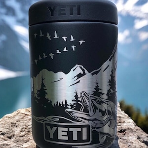 K&ouml;nnte beinhalten: Schwarzer YETI-Dosenk&uuml;hler mit Berg- und Fischdesign. Das Design ist in Silber ge&auml;tzt. Der K&uuml;hler hat einen schwarzen Deckel und das YETI-Logo ist auf der Vorderseite abgebildet.