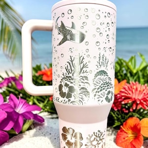 May include: A light pink insulated tumbler with a handle, lid, and straw. The tumbler features a silver design of a whale shark, coral, and bubbles. The background shows a beach scene with flowers.
