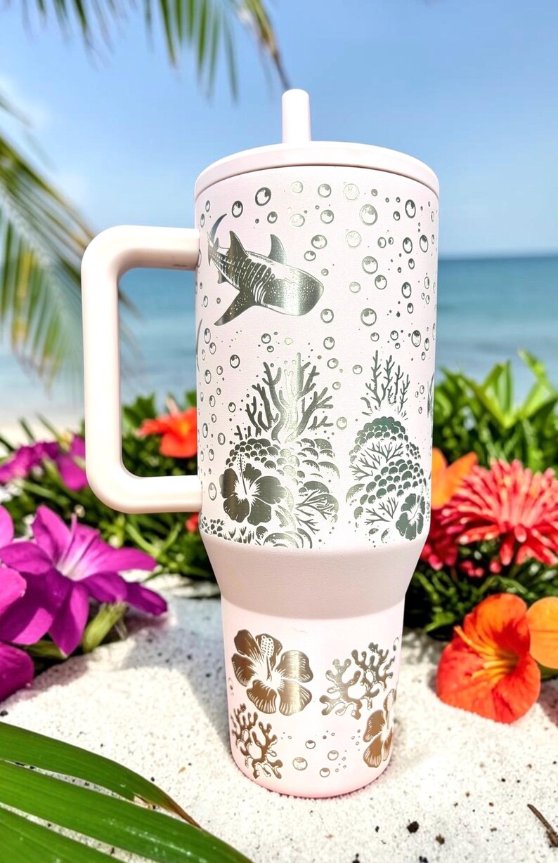 May include: A light pink insulated tumbler with a handle, lid, and straw. The tumbler features a silver design of a whale shark, coral, and bubbles. The background shows a beach scene with flowers.