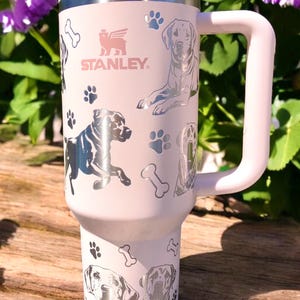 Peut inclure: Une tasse de voyage en acier inoxydable rose avec une poign&eacute;e noire. La tasse pr&eacute;sente un motif argent&eacute; de chiens et d'empreintes de pattes. La tasse porte le texte "STANLEY" sur le devant.