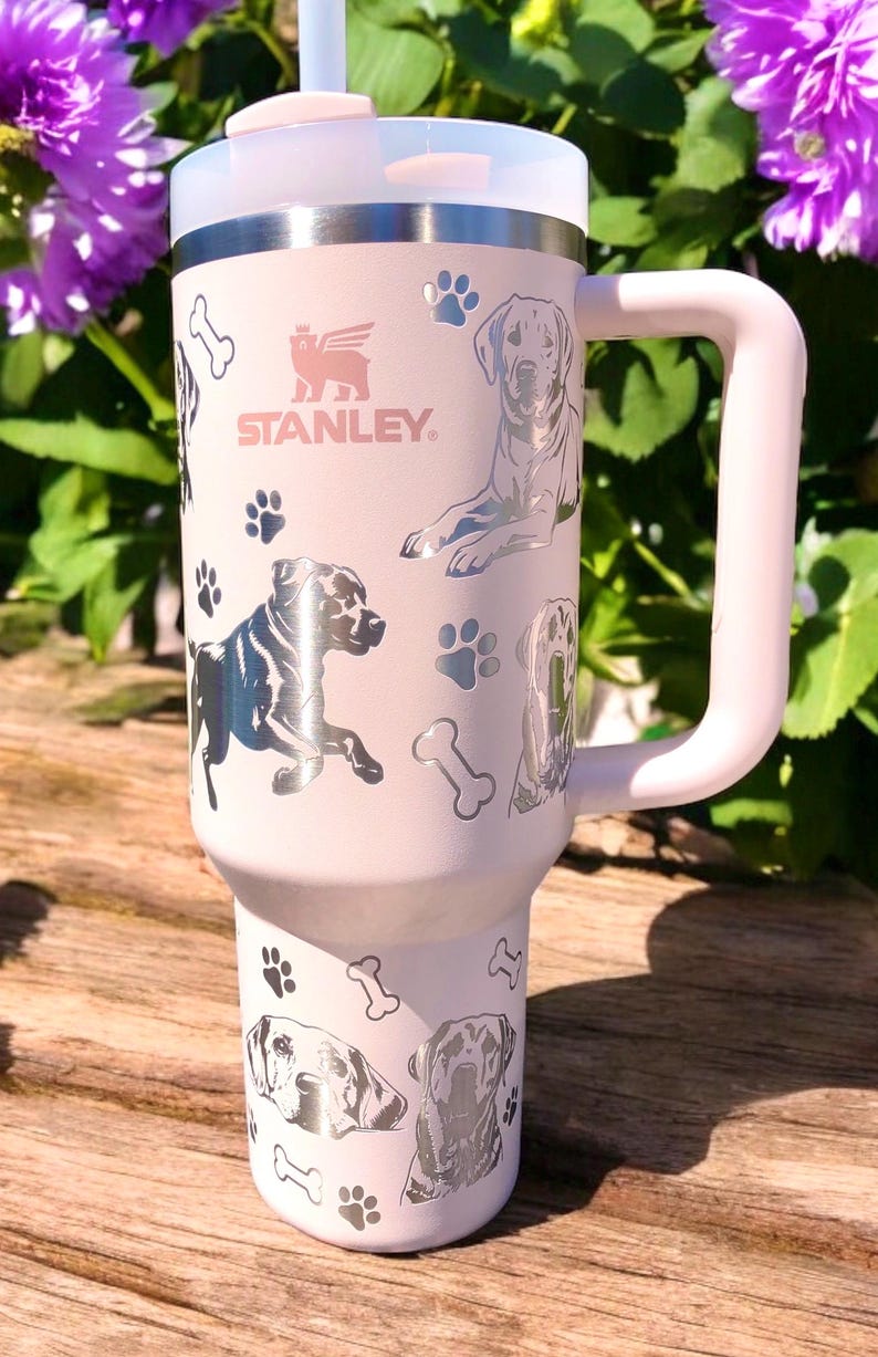 Peut inclure: Une tasse de voyage en acier inoxydable rose avec une poign&eacute;e noire. La tasse pr&eacute;sente un motif argent&eacute; de chiens et d'empreintes de pattes. La tasse porte le texte "STANLEY" sur le devant.