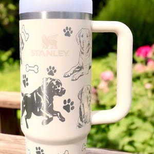 Peut inclure: Mug de voyage en acier inoxydable blanc avec une poign&eacute;e noire et une paille. Le mug est d&eacute;cor&eacute; d'empreintes de pattes de chien et d'os avec le mot "STANLEY" imprim&eacute; sur le devant.