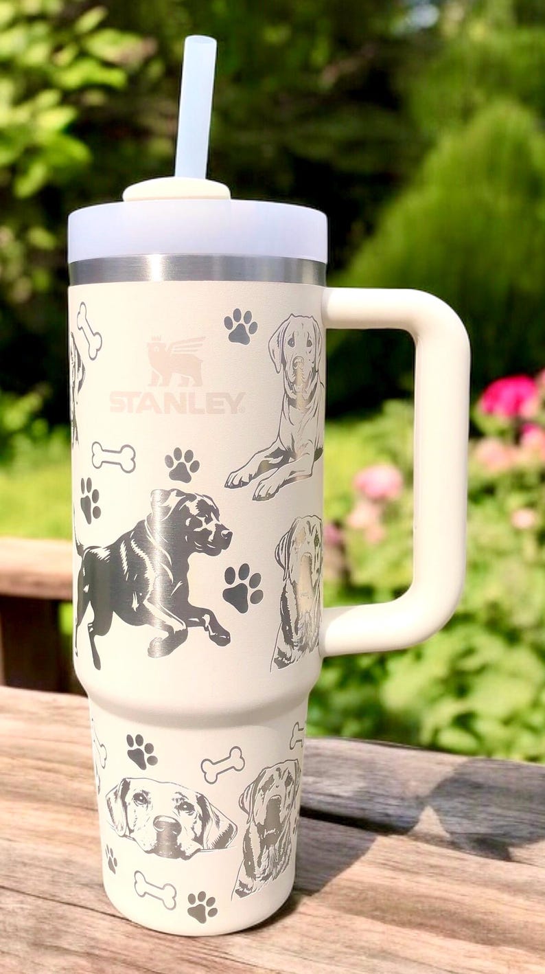 Peut inclure: Mug de voyage en acier inoxydable blanc avec une poign&eacute;e noire et une paille. Le mug est d&eacute;cor&eacute; d'empreintes de pattes de chien et d'os avec le mot "STANLEY" imprim&eacute; sur le devant.