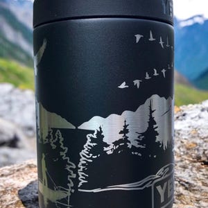 K&ouml;nnte beinhalten: Schwarzer isolierter Becher mit einer silberfarbenen, ge&auml;tzten Au&szlig;enszene mit B&auml;umen, V&ouml;geln und einer angelnden Person. Der Becher hat einen schwarzen Deckel und die Marke "YETI" ist sichtbar.