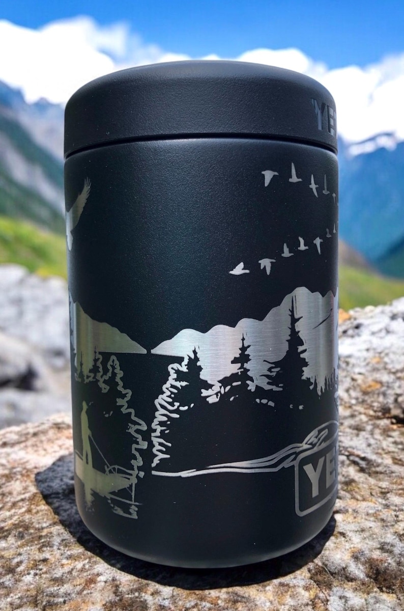 K&ouml;nnte beinhalten: Schwarzer isolierter Becher mit einer silberfarbenen, ge&auml;tzten Au&szlig;enszene mit B&auml;umen, V&ouml;geln und einer angelnden Person. Der Becher hat einen schwarzen Deckel und die Marke "YETI" ist sichtbar.