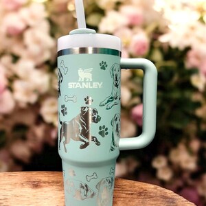 Peut inclure: Une tasse de voyage en acier inoxydable bleu clair avec une poign&eacute;e et une paille. La tasse est orn&eacute;e d'un motif argent&eacute; d'empreintes de pattes de chien et du mot "STANLEY" imprim&eacute; sur le c&ocirc;t&eacute;.