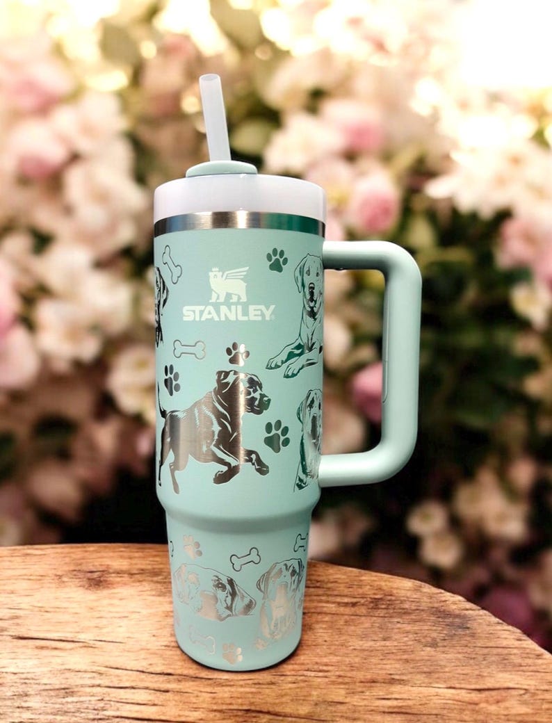 Peut inclure: Une tasse de voyage en acier inoxydable bleu clair avec une poign&eacute;e et une paille. La tasse est orn&eacute;e d'un motif argent&eacute; d'empreintes de pattes de chien et du mot "STANLEY" imprim&eacute; sur le c&ocirc;t&eacute;.