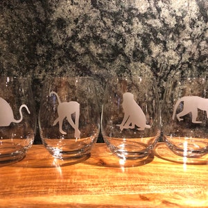 May include: Four clear glass wine glasses with etched silhouettes of monkeys and a leopard. The glasses are arranged in a row on a wooden surface.
