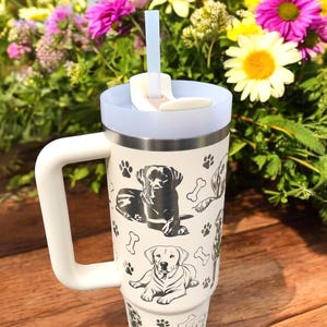 Peut inclure: Mug de voyage en acier inoxydable blanc avec une poign&eacute;e noire et un motif d'empreintes de pattes de chien noir et blanc. Le mug a un couvercle avec un trou pour paille et une paille.