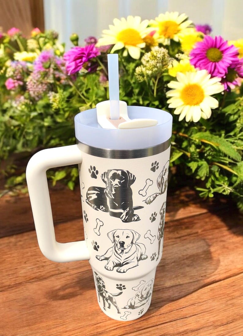 Peut inclure: Mug de voyage en acier inoxydable blanc avec une poign&eacute;e noire et un motif d'empreintes de pattes de chien noir et blanc. Le mug a un couvercle avec un trou pour paille et une paille.