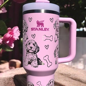May include: A pink stainless steel travel mug with a black and white doodle pattern of dogs, bones, and hearts. The mug has a handle and a lid with a straw. The mug has the text "STANLEY" on it.
