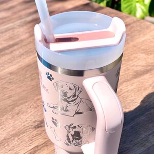 Peut inclure: Une tasse &agrave; caf&eacute; isotherme en acier inoxydable rose avec un couvercle transparent et une paille. La tasse a une poign&eacute;e argent&eacute;e et un motif argent&eacute; de chiens et de pattes.