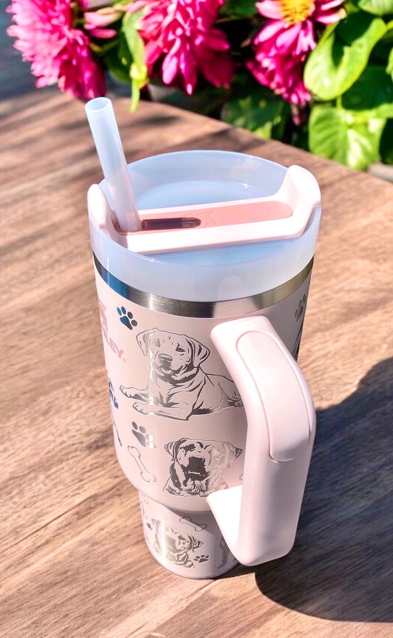 Peut inclure: Une tasse &agrave; caf&eacute; isotherme en acier inoxydable rose avec un couvercle transparent et une paille. La tasse a une poign&eacute;e argent&eacute;e et un motif argent&eacute; de chiens et de pattes.