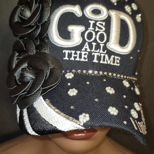 May include: A black baseball cap featuring the embroidered white text GOD IS ALL THE TIME. The cap is decorated with black leather-like floral appliques on the left side, white faux pearls, and small clear rhinestones across the front and brim.
