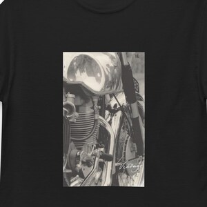 Könnte beinhalten: Schwarzes T-Shirt mit einem Sepia-Ton-Bild eines Oldtimer-Motorrads. Das Bild zeigt die Chromdetails des Motorrads, einschließlich des Kraftstofftanks und des Vorderrads. Das T-Shirt hängt an einem schwarzen Kleiderbügel.