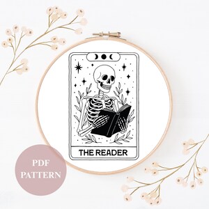 Könnte beinhalten: Ein schwarz-weißer Stickrahmen mit einem Tarotkarten-Design. Die Karte zeigt ein Skelett, das ein Buch liest, mit den Worten "THE READER" unten. Das Design umfasst Sterne, Blätter und Mondphasen. Ein rosa Kreis mit "PDF PATTERN" befindet sich unten links.