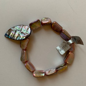 Op de afbeelding: Een roze en iriserende schelpenarmband met een grote iriserende schelpenhanger. De armband heeft een label met de tekst "LooLoo Kids" en "€ 10".