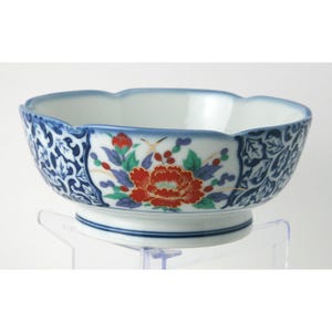 May include: A white ceramic bowl with a scalloped rim, featuring a central red peony floral motif with green leaves and blue accents. The exterior is decorated with a dense blue and white scrolling leaf pattern on both sides of the floral panel.