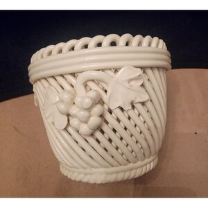 May include: A white ceramic basket with a woven design. The basket features a decorative grape cluster and leaf motif on the front. The rim has a looped design. The basket is likely used for decorative purposes.