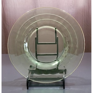 May include: A clear, pale green glass plate with a gold rim, presented on a dark green metal stand. The plate features concentric rings and a textured design near the centre. The background is a muted pink and grey.