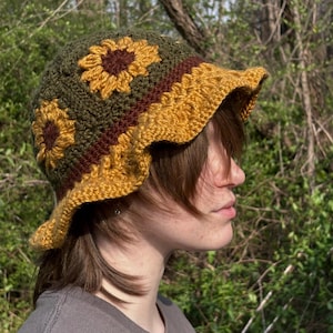 May include: A handmade crochet bucket hat featuring a sunflower design. The hat has a green base with yellow and brown sunflower appliques and a yellow scalloped brim. The hat is a unique, stylish accessory.