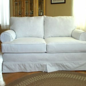 Könnte beinhalten: Ein weißes Zweisitzer-Sofa mit einem gerüschten Sockel und gerollten Armlehnen. Das Sofa hat zwei große Rückenkissen und zwei Sitzkissen. Der Raum hat Holzböden und einen runden Teppich.