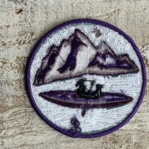 Op de afbeelding: Ronde geborduurde patch met een bergketen en een kajak op een meer, in tinten paars, grijs en wit. De patch heeft een paarse rand en is waarschijnlijk voor een jas of rugzak.