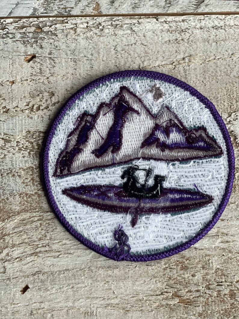 Op de afbeelding: Ronde geborduurde patch met een bergketen en een kajak op een meer, in tinten paars, grijs en wit. De patch heeft een paarse rand en is waarschijnlijk voor een jas of rugzak.